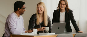 Three people having a discussion around a table with a laptop, notebook, and coffee cups.