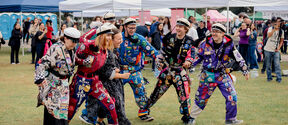 A group of students in colourful overalls decorated with patches, standing on a lawn at an outdoor event with tents.