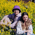 Two Aalto University students sitting outdoors in a field of yellow flowers, one wearing a purple hat and the other in a white jacket.