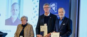 Kolme henkilöä pitelemässä todistuksia palkintotilaisuudessa. Taustalla sininen banneri ja iso näyttö.