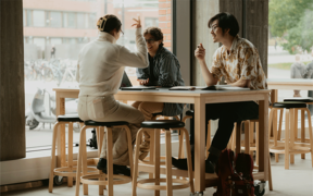 Three people sitting at a wooden table in a modern space, engaged in a discussion with a laptop.