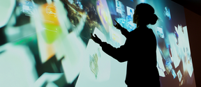 AI generated vision of a teacher interacting with virtual screens in a dark room, with colourful digital images projected on the walls.
